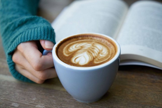Hånd holder kaffekopp med hjertemønster i kaffen. Bok i bakgrunnen.