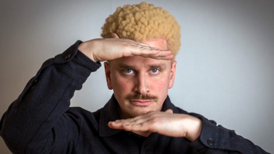 En alvorlig mann med bart og lys afro lager et vindu med henendene og titter igjennom