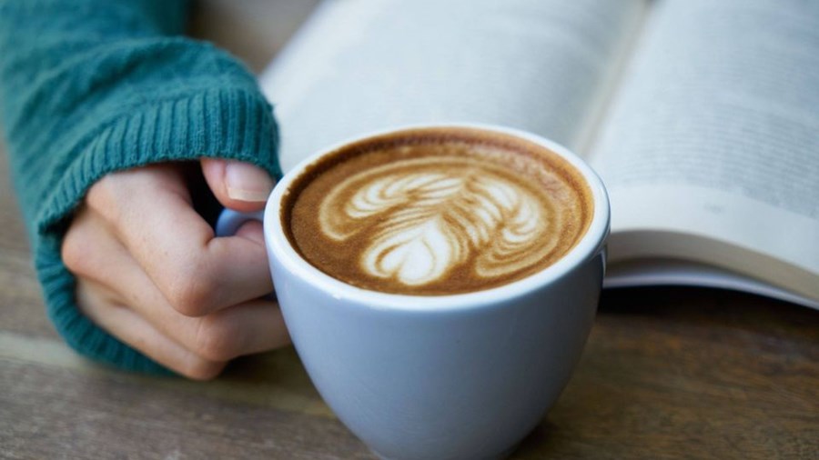 Hånd holder kaffekopp med hjertemønster i kaffen. Bok i bakgrunnen.